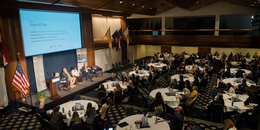an aerial view of a large meeting room with people at tables watching a presentation on stage