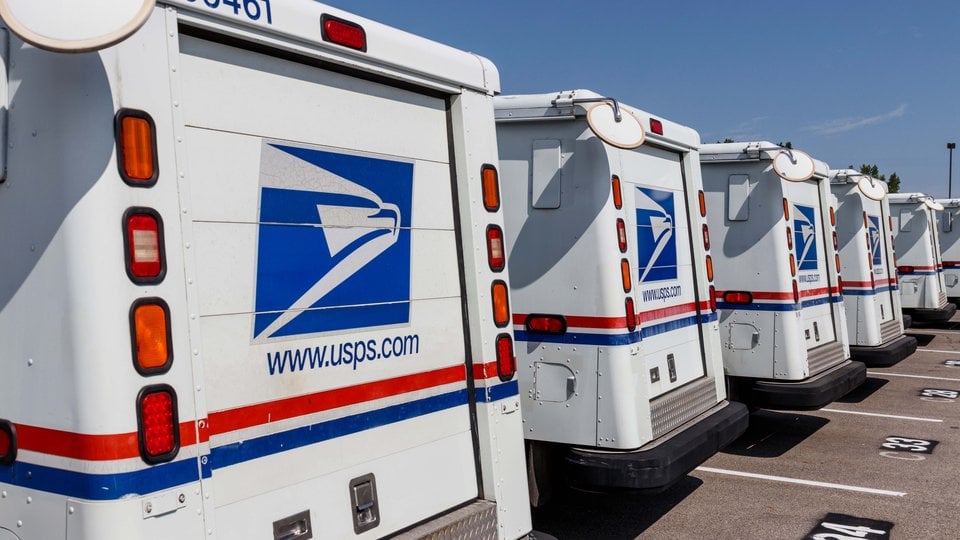 A row of US Postal trucks lined up in a parking lot
