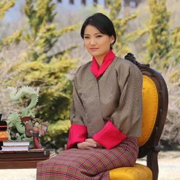 The Queen of Bhutan sits in an ornate wooden chair with yellow cushions and smiles gently. She is wearing traditional Bhutanese clothing.
