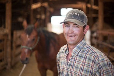 A man with a plaid shirt and baseball cap smiles in a barn in front of a horse