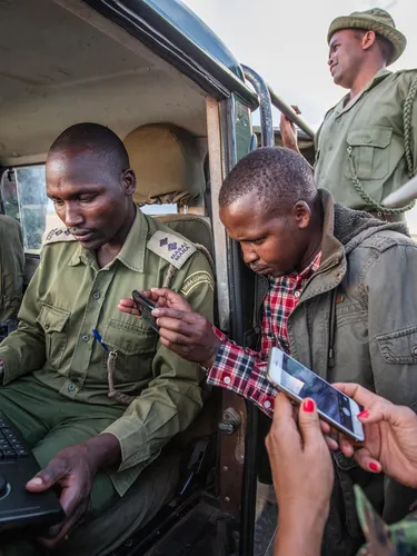 Group of people stand around an open jeep looking at a hand-held device with keyboard