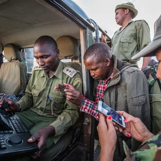 Group of people stand around an open jeep looking at a hand-held device with keyboard