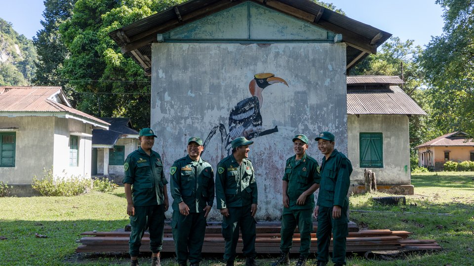 Men in uniform stand in front of building with mural of a toucan