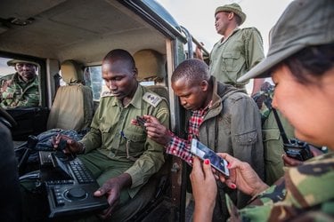 Group of people stand around an open jeep looking at a hand-held device with keyboard