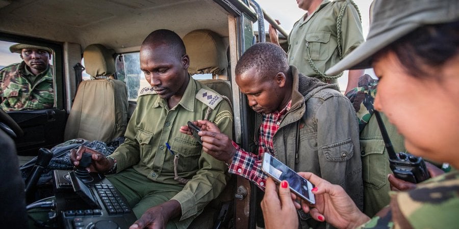Group of people stand around an open jeep looking at a hand-held device with keyboard