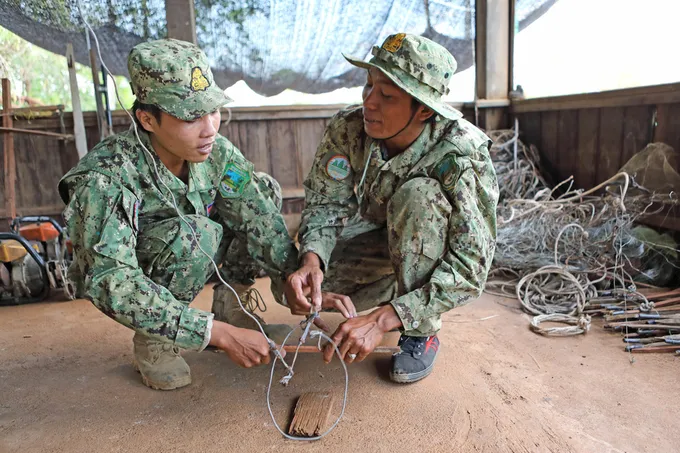 Two rangers dressed in green camouflage demonstrate a snare with a loop of rope and a stick