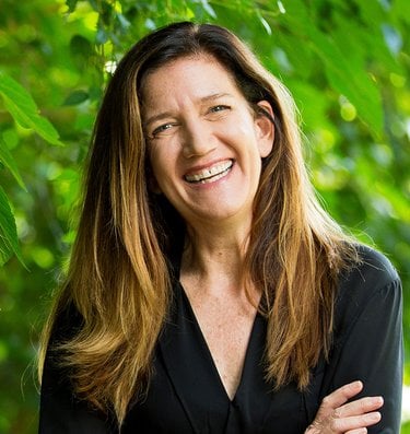 A photo of a woman with long medium brown hair wearing a black v-nek top and smiling at the camera with a green background