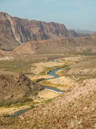 A color photo with brown mountains in the distance, and a dry valley with patches of green grass and a river flowing in the center.