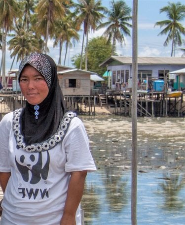 A portrait of a woman standing on a dock wearing a black headdress and white tshirt with a village on stilts in the background on the shoreline