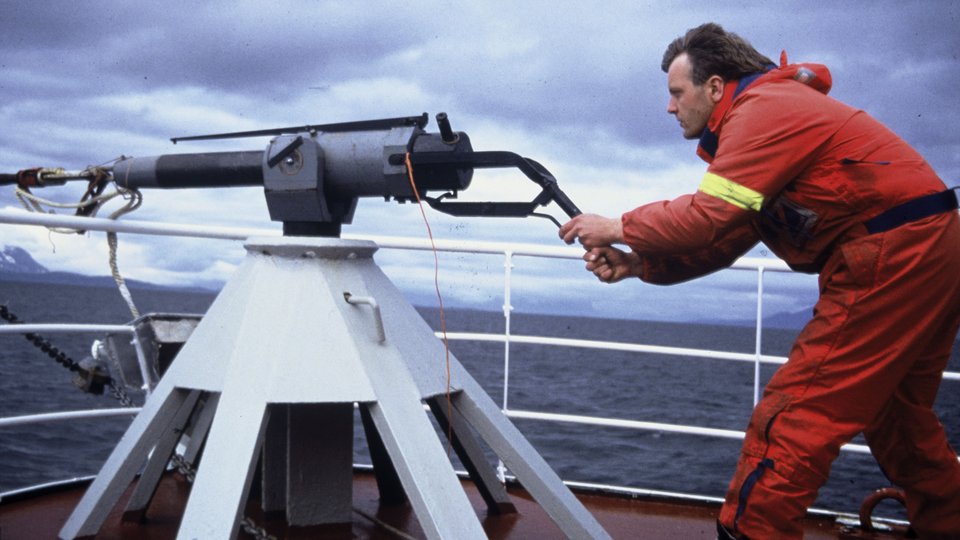 A man on a ship's deck aims a large harpoon gun
