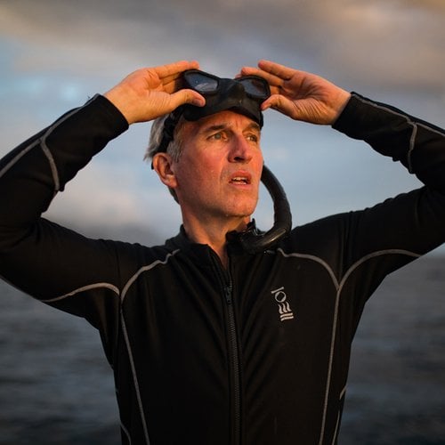 A headshot of a man standing next to the ocean with snorkel goggles on his forehead