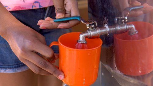 A hand holds a red, plastic mug under a spigot which is turned on with the other hand