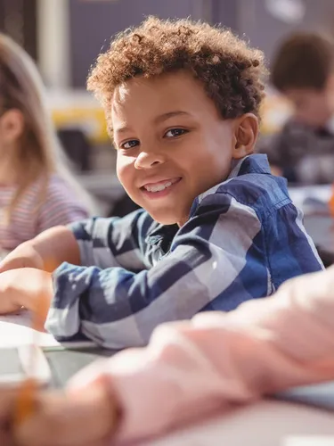 A Black boy sits in a classroom with his fellow classmates. He looks at the camera and smiles.