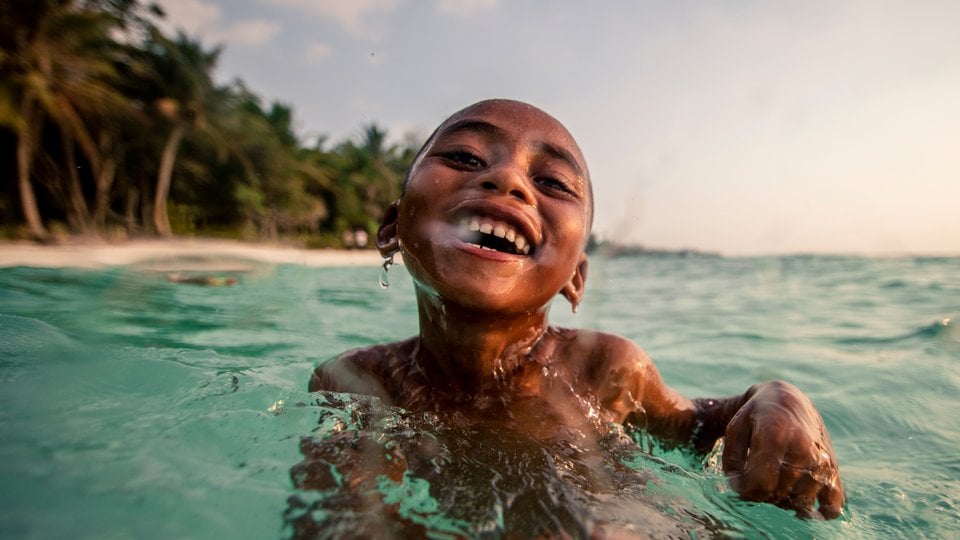 A close up image of a smiling kid swimming in turquoise blue water with palms in the background.
