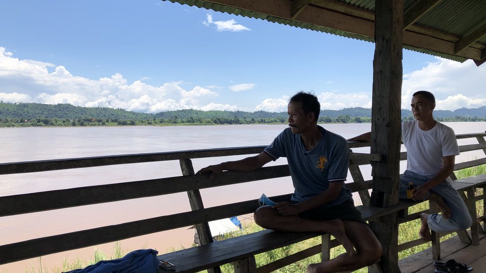 Two men sit on a wooden bench under a gazeebo looking out over a river below