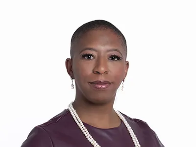 Headshot of a Black woman with a shaved head. She is wearing a pleather maroon shirt and a string of pearls.