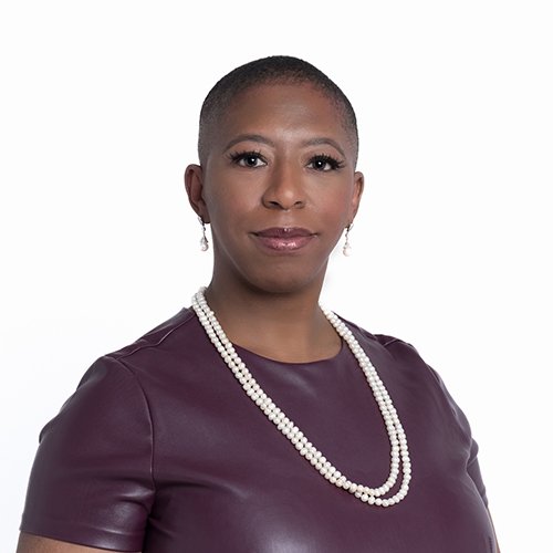 Headshot of a Black woman with a shaved head. She is wearing a pleather maroon shirt and a string of pearls.