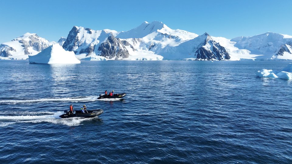 Two small zodiac boats cruise through icy blue waters with icebergs and snow covered Antarctic mountains in the background.