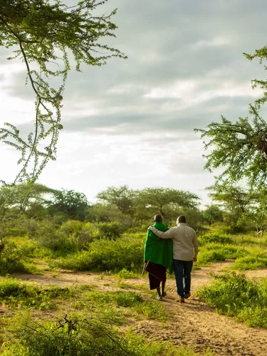 Photo of Two Tanzanian men walking side-by-side away from the camera and into the beautiful landscape. The man on the right has his arm around the others' shoulder.
