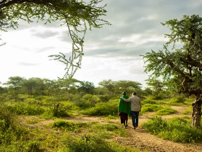 Photo of Two Tanzanian men walking side-by-side away from the camera and into the beautiful landscape. The man on the right has his arm around the others' shoulder.