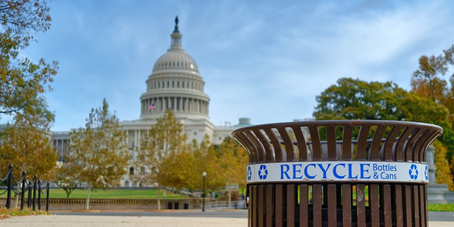 A color photo showing the Capitol building in Washington DC with a trash can with a recycle label in the foreground with trees with yellow leaves in the mid-distance
