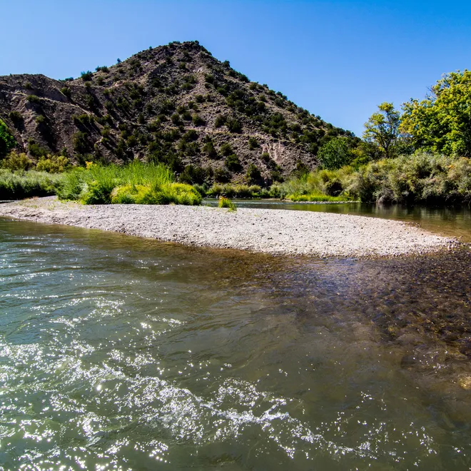 A color photo showing the Rio Grande River in the foreground with a sandy spit in the middle and a small mountain and trees in the background