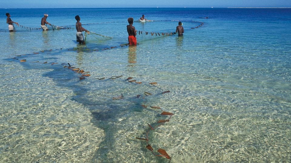 A group of six fishermen wade in the calm water with a large net to catch some fish.