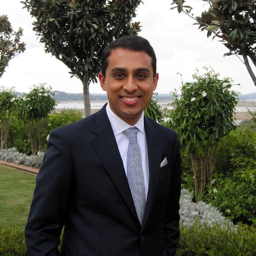 Vignesh Aier smiles at the camera, wearing a suit with his hands in his pockets with greenery behind him