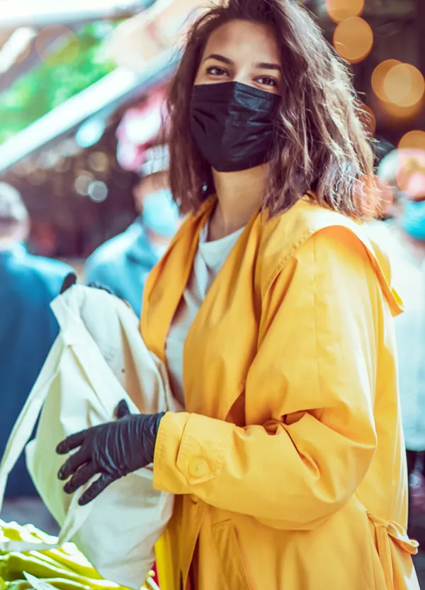 A photo of woman wearing a yellow jacket, black gloves, and a black mask over her mouth holding a bag.