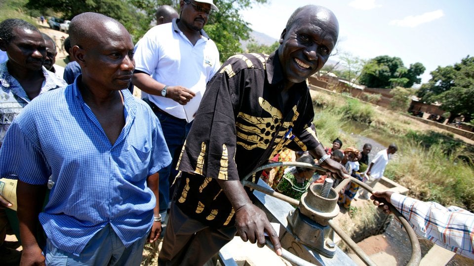 A group of men smile as one turns a giant wheel on a water pump