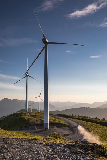 Several windmills on grassy hills. A car drives on a gravel road below the windmills, showing their massive scale.