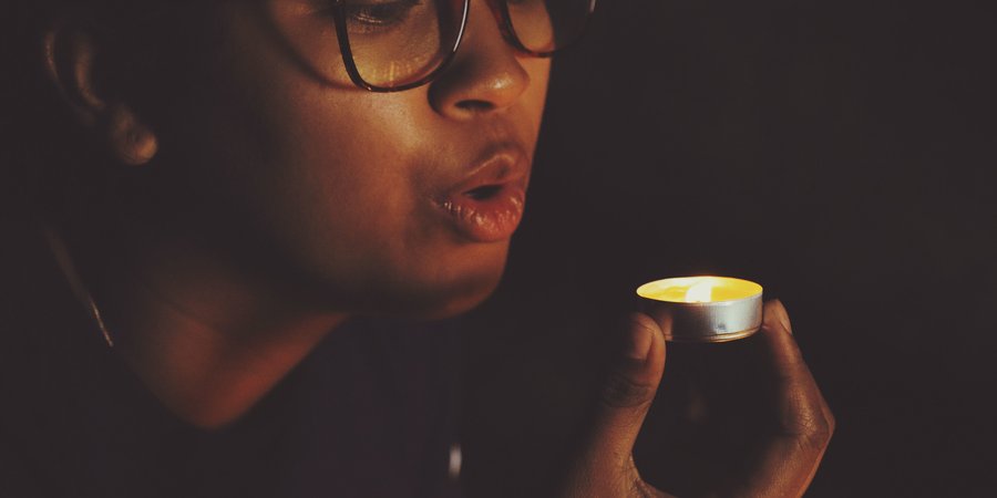 A person wearing glasses in the dark blowing out a small candle.