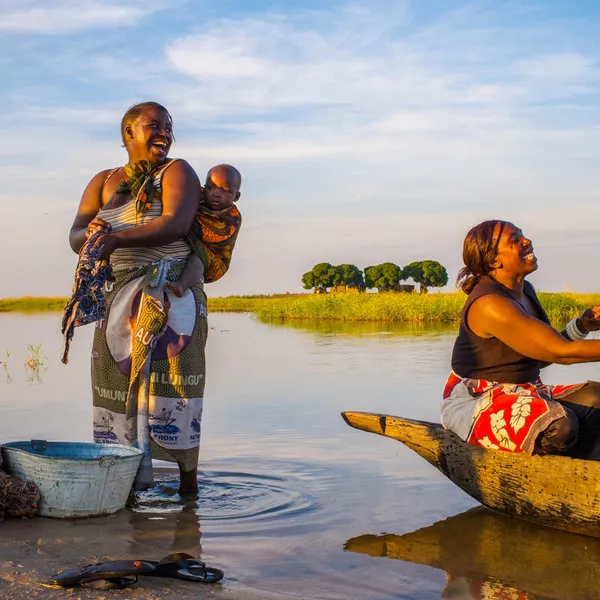 Two women and a baby on a river bank, one washing clothes and the other casting a net while all smile