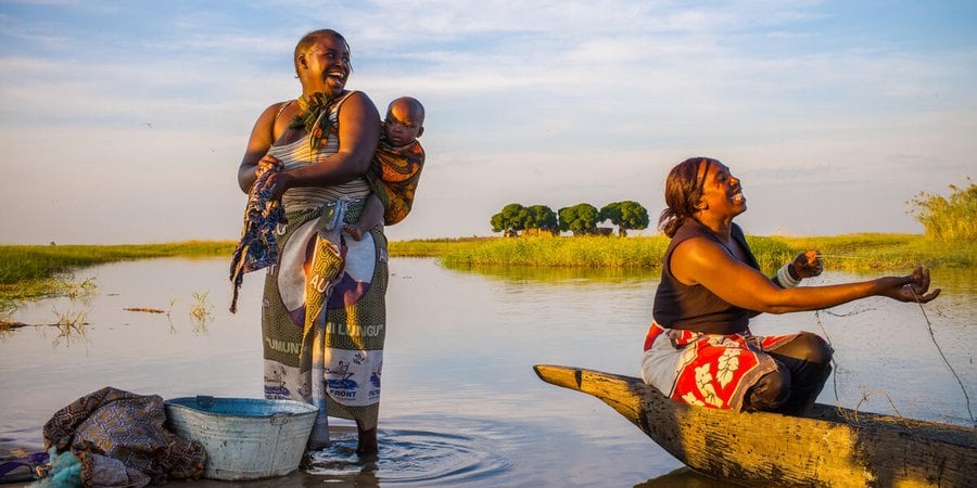 Two women and a baby on a river bank, one washing clothes and the other casting a net while all smile