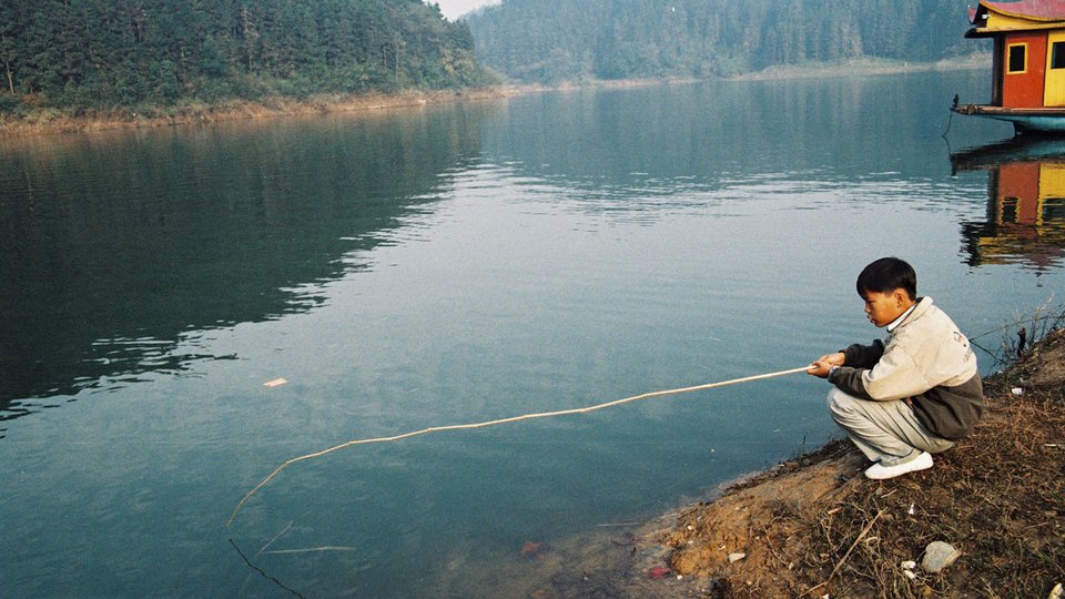 A man holding a fishing rod on a brown shoreline with a dark river and small tree-covered hills in the background.