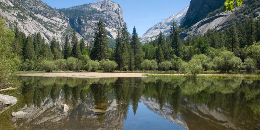 A photo of mountains and a trees reflecting on a waterway with blue skies in the backaground
