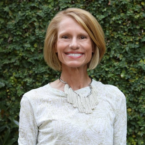 A color photo of a white woman with a bob haircut, a white shirt and necklace, smiling at the camera