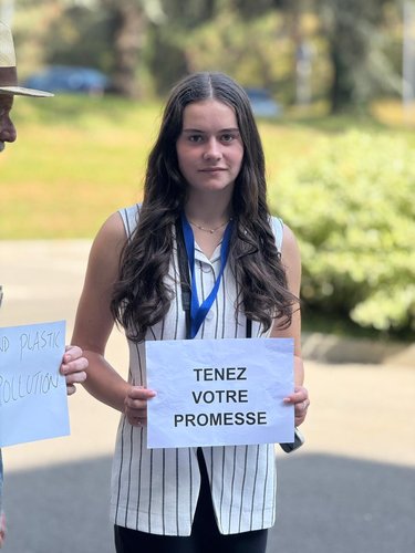 A woman with long brown hair holding a sign reading "Tenez Votre Promesse" outisde