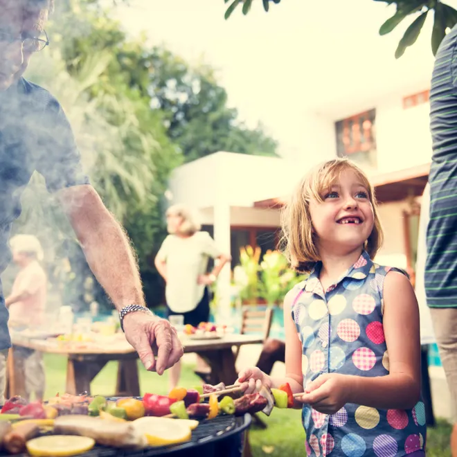 A summer cookout scene of a child smiling and helping to barbecue some veggie skewers with the supervision of two adults.