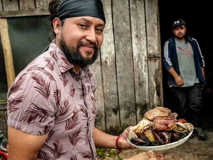 Member of the Huilliche Indigenous community in Yaldad, Chile, looks to camera as he walks with two plates of curanto, a traditional dish
