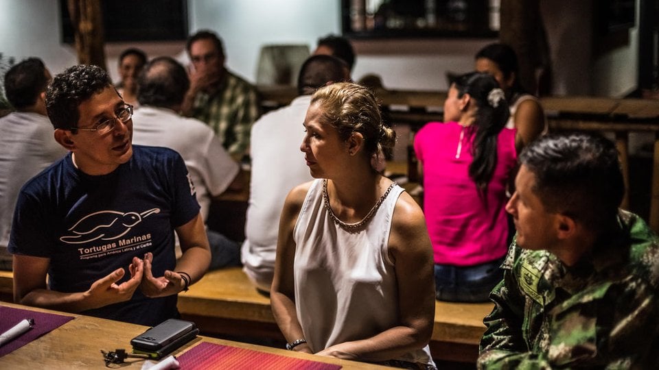 A woman sitting between two men at a dinner table in conversation with additional people behind them at other tables
