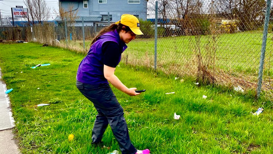 Woman in a yellow hat standing in the grass next to a chain link fence taking photos with a cell phone of trash in the grass