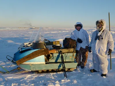 A man and a snowmobile standing in front of a snowy mountain range with another person walking down the mountain behind him