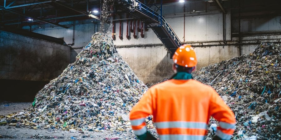 Rear view of young male worker in helmet, pollution mask, and reflective clothing observing waste falling from conveyor belt onto pile at facility.