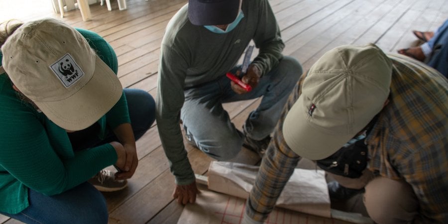 A color photo of three people wearing baseball caps inside looking down at some planning documents on a wooden floor.