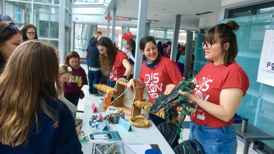 three women in red tshirts standing behind white folding tables showing passersby their educational materials