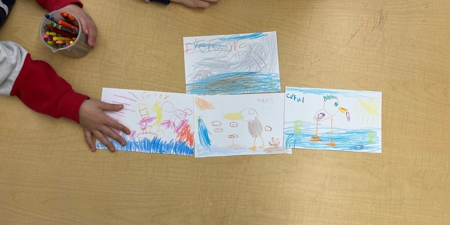 A color photo of a wooden table with children's arms reaching out to four papers with children's colorful drawings