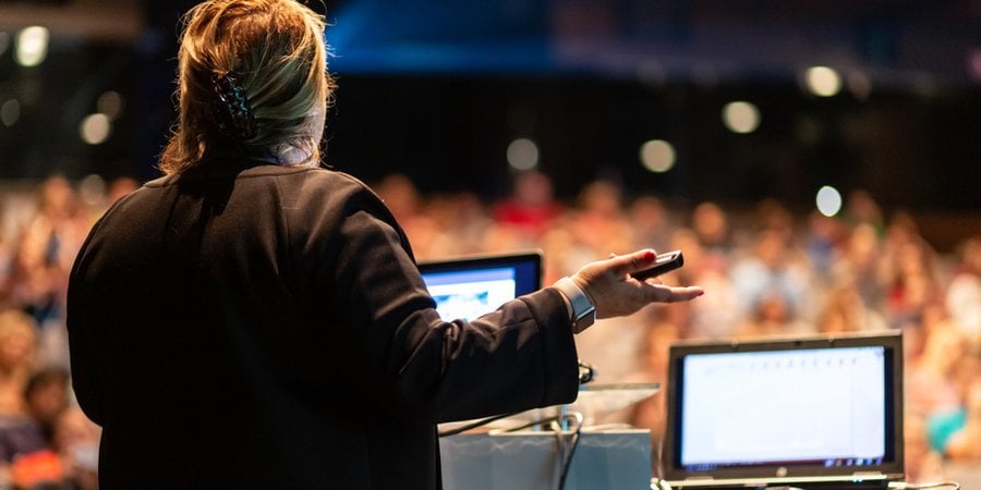 A speaker with her back to the camera addresses a crowd