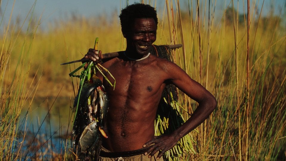 A fisherman stands in tall reeds holding his catch over his shoulder with a hand on his hip